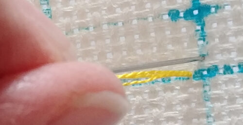 A close up on a needle with yellow floss being inserted into white, gridded aida fabric