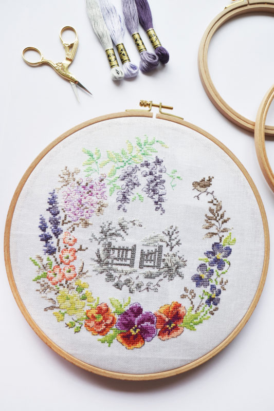 A cross stitch of a floral border with a back and white cottage in the middle
