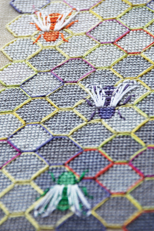 A close up on a stitched honey comb pattern with cross stitch bees on a blue-grey aida background