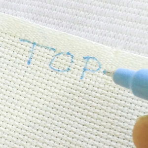 Close up on a serged piece of aida fabric with the word Top written clearly across the top in blue ink