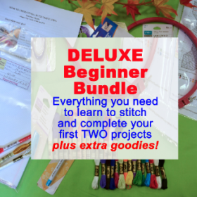 A green tabletop, bordered by a strand of fall leaves across the top. It holds an embroidery pattern, a large set of floss, a hoop, various tools, and samples of premium fabrics. Text reads: "DELUXE Beginner Bundle, Everything you need to learn to stitch and complete your first TWO projects, plus extra goodies!"