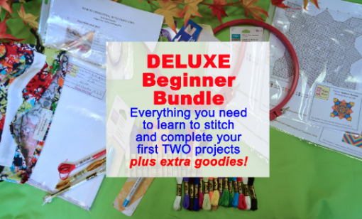 A green tabletop, bordered by a strand of fall leaves across the top. It holds an embroidery pattern, a large set of floss, a hoop, various tools, and samples of premium fabrics. Text reads: "DELUXE Beginner Bundle, Everything you need to learn to stitch and complete your first TWO projects, plus extra goodies!"